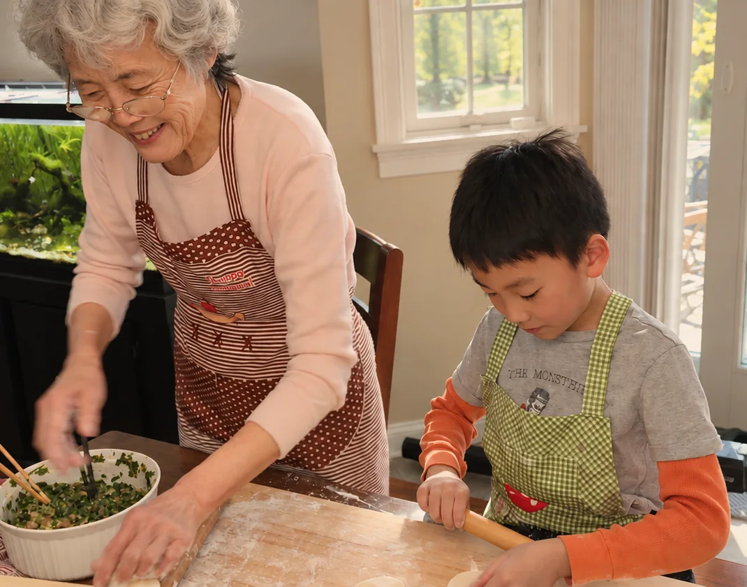 Young Ethan in a green checked apron rolling dumpling wrappers at a floured board while a family member in a polka-dot apron mixes a green filling beside him