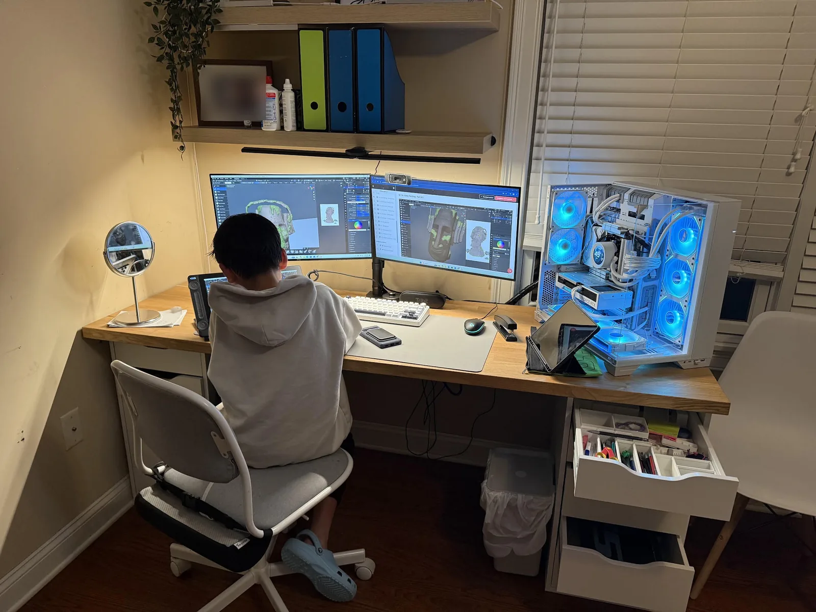 Ethan seen from behind at his desk, working on a 3D model of a sculpted head across two monitors, next to a computer with glowing blue fans