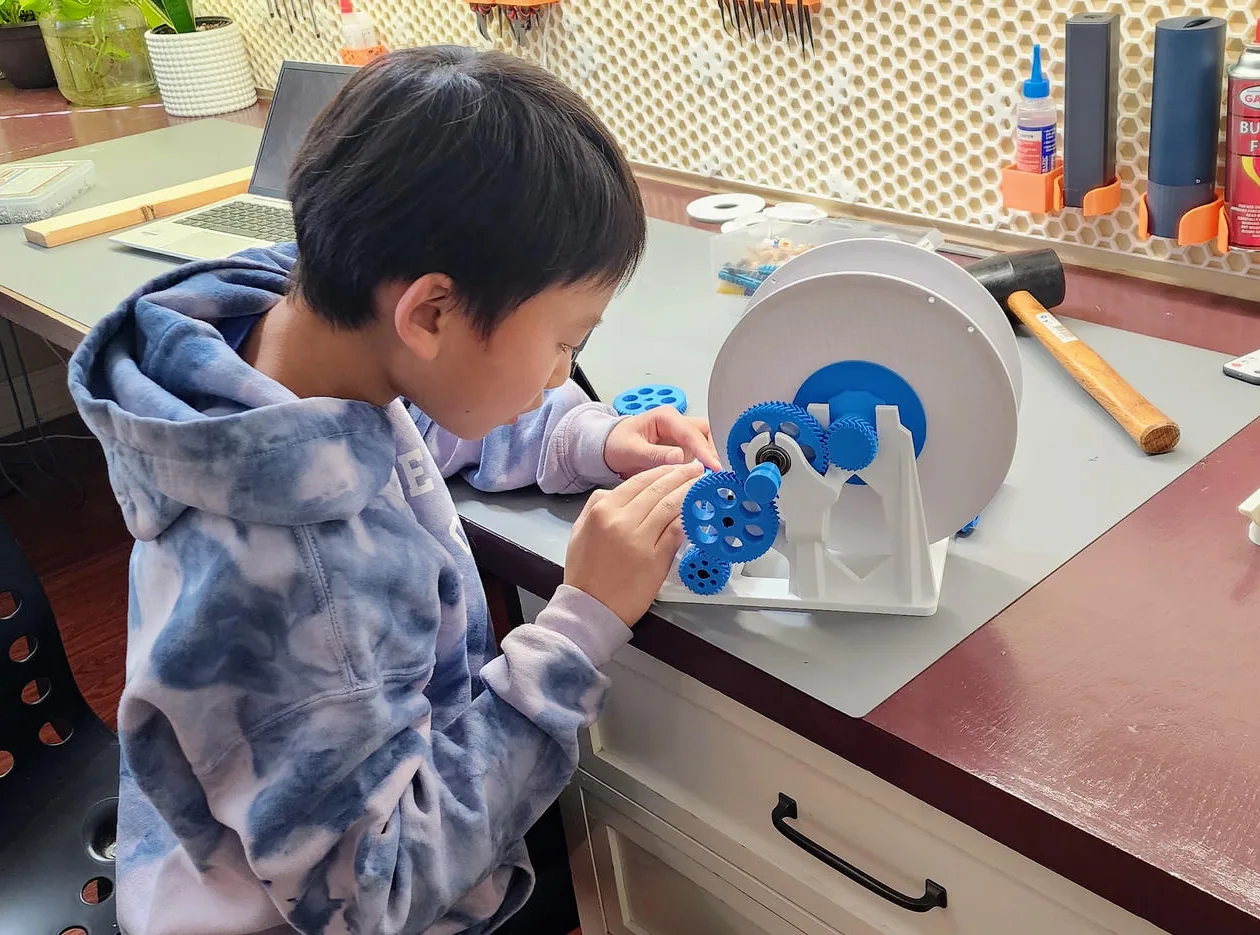 Ethan in a blue tie-dye hoodie fitting blue 3D-printed gears onto a white printed spool holder at his workbench, with tools on a honeycomb pegboard behind him