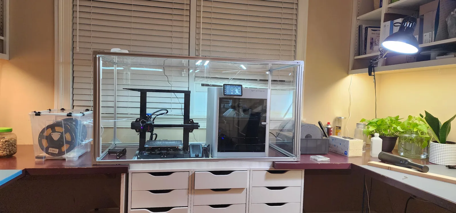 Ethan's home studio: two 3D printers inside a large clear enclosure on a desk with white drawers, a filament box on the left, and plants under a desk lamp on the right